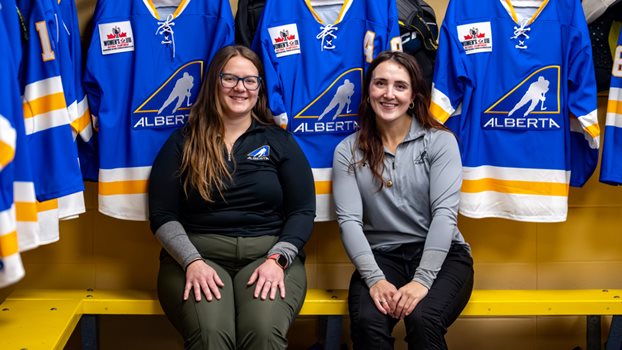 Jessie Olfert (left) and Jane Kish sit on the bench of Team Alberta's locker room, with team jerseys hanging in the stalls behind them.